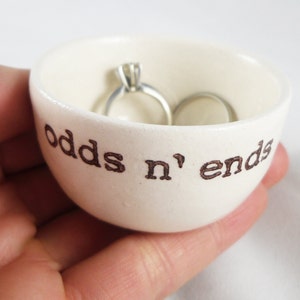 May include: White ceramic ring dish with the words "odds n' ends" inscribed on the front. Two silver rings are resting inside the dish.