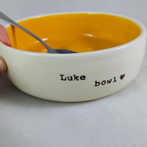 May include: A white ceramic bowl with a yellow interior. The bowl has the words "Luke bowl" and a heart symbol written on the outside.