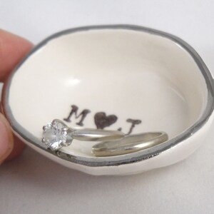 May include: White ceramic ring dish with a silver rim. The dish has the initials "M & J" with a heart in between. A diamond engagement ring is resting in the dish.