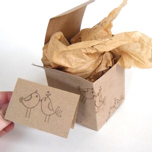 May include: A brown cardboard box with a lid, filled with brown tissue paper. The box is decorated with a repeating pattern of hand-drawn birds. A small brown card with a hand-drawn image of two birds kissing is placed in front of the box.