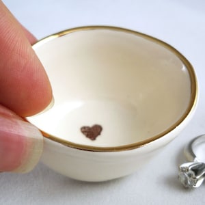 May include: A small white ceramic ring dish with a gold rim and a brown heart design in the center.