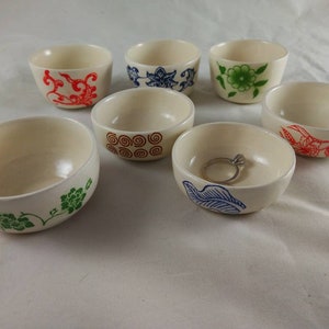 May include: Set of seven small white ceramic bowls with various colorful floral and geometric designs. The bowls are perfect for holding small items like jewelry or trinkets.
