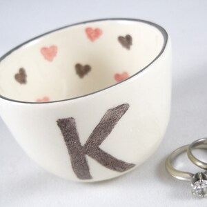 May include: White ceramic ring dish with a brown letter K and pink and brown hearts. The dish is filled with two silver wedding bands.