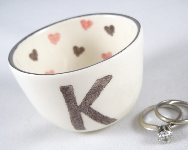 May include: White ceramic ring dish with a brown letter K and pink and brown hearts. The dish is filled with two silver wedding bands.