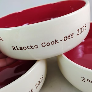 Puede incluir: Tres cuencos de cerámica blanca con interior rojo. Los cuencos están apilados uno encima del otro. El cuenco superior tiene el texto "1er Risotto Cook-Off 2025" escrito en él. El cuenco del medio tiene el texto "2025" escrito en él. El cuenco inferior tiene el texto "2º" escrito en él.