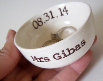May include: White ceramic ring dish with the text "08.31.14" and "Mrs. Gibas" in brown lettering. Two silver rings are inside the dish.
