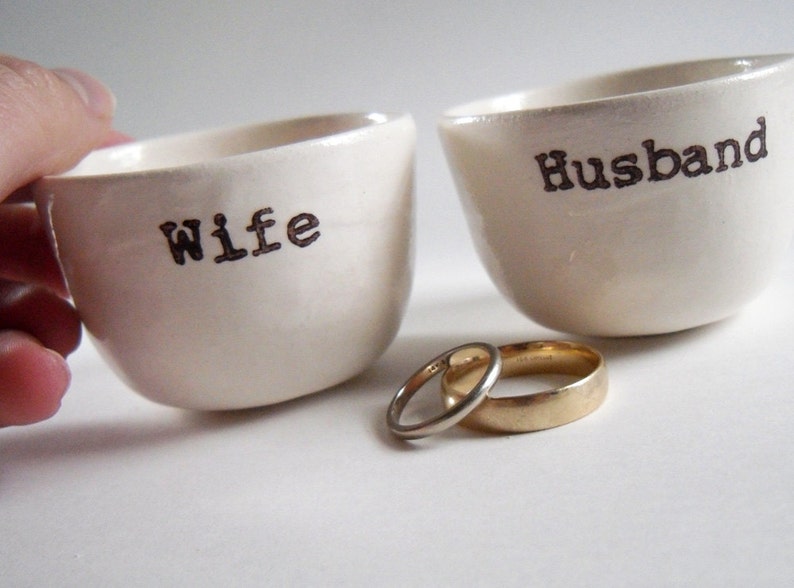 Puede incluir: Dos cuencos de cer&aacute;mica blanca con las palabras "Wife" y "Husband" impresas en ellos. Dos alianzas de oro se colocan delante de los cuencos.