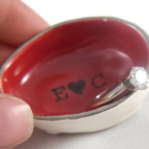 Pode incluir: Um prato de anel de cerâmica vermelha com uma borda branca e a inscrição "E & C" com um coração entre as letras. Um anel de noivado de diamante está descansando no prato.