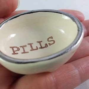 May include: A small white ceramic dish with a silver rim. The dish has the word "PILLS" printed in brown lettering on the inside.