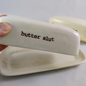 May include: Off-white ceramic butter dish with the words "butter slut" printed in brown. The dish has a rectangular base and a matching lid. Another butter dish is visible in the background.