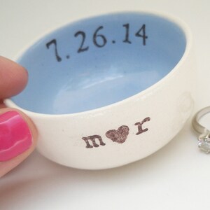 Puede incluir: Un plato de anillo de cerámica blanca con un interior azul claro. El plato está inscrito con la fecha "7.26.14" y las iniciales "M & R" con un símbolo de corazón entre las letras.