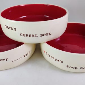May include: Three ceramic bowls with red interiors and white exteriors. The bowls have different inscriptions: "Papa's Cereal Bowl", "U.S. Army --- Eric", and "Grandpa's Soup Bowl".