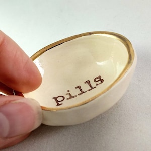 Puede incluir: Un pequeño plato de cerámica blanca con un borde dorado. El plato tiene la palabra "pills" escrita en el interior en letras marrones.