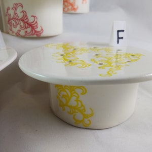 May include: White ceramic planter with a yellow floral design. The planter has drainage holes and a matching saucer. The planter is labeled with the letter "F".