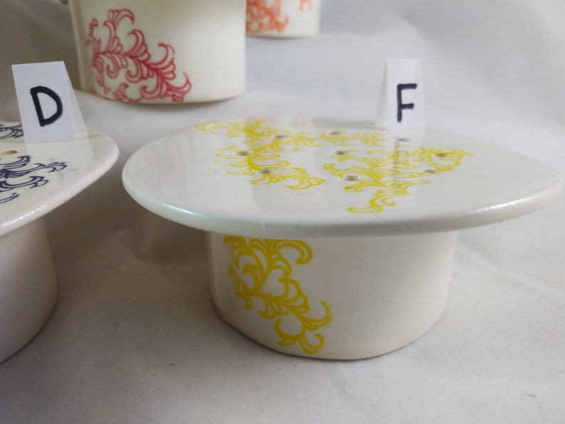 May include: White ceramic planter with a yellow floral design. The planter has drainage holes and a matching saucer. The planter is labeled with the letter "F".