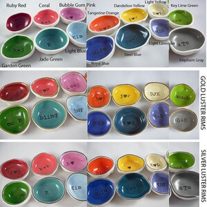 May include: A collection of small, irregularly shaped ceramic ring dishes in various colors, including red, pink, yellow, and blue. Each dish has a white interior and a gold or silver luster rim, with text such as initials or words like "Yes!" or "Bling".