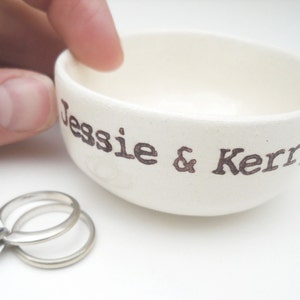 May include: White ceramic ring dish with the names "Jessie & Kerry" written in brown lettering. A silver ring with a diamond is sitting on the dish.