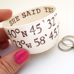 May include: A white ceramic ring dish with the words "SHE SAID YES" and coordinates printed in brown. Two silver rings with a diamond are next to the dish. The dish is held by a person with pink nail polish.