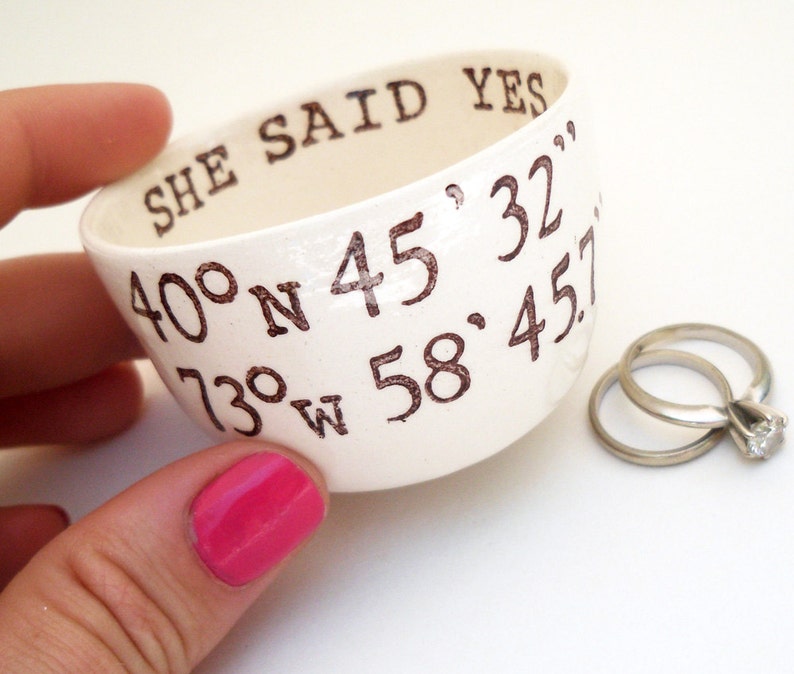 May include: A white ceramic ring dish with the words "SHE SAID YES" and coordinates printed in brown. Two silver rings with a diamond are next to the dish. The dish is held by a person with pink nail polish.