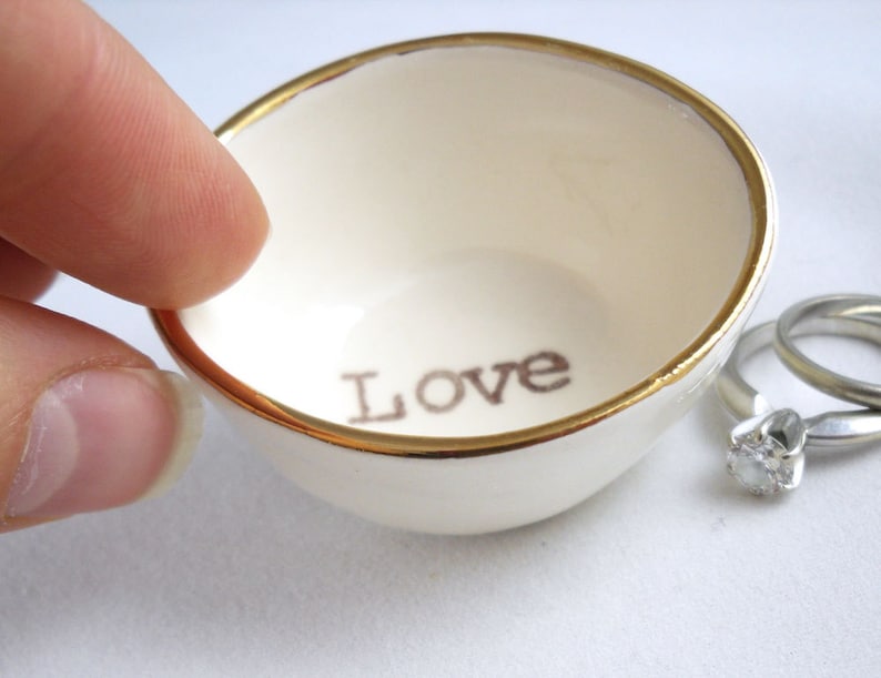 May include: White ceramic ring dish with a gold rim and the word "Love" inscribed on the inside. Two silver rings are sitting next to the dish.