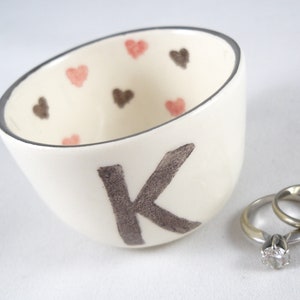May include: White ceramic ring dish with a gray letter K and pink and brown hearts. Two silver rings are in the dish.