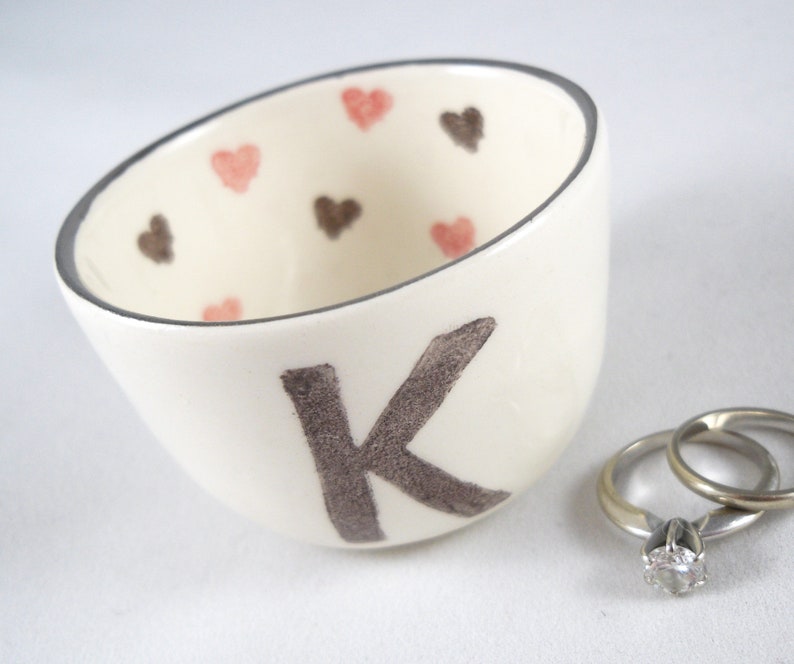 May include: White ceramic ring dish with a gray letter K and pink and brown hearts. Two silver rings are in the dish.
