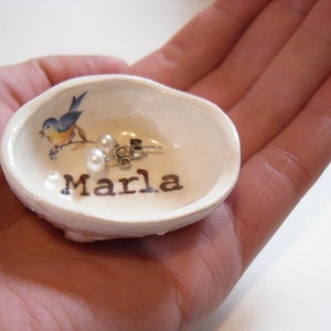 May include: A white ceramic dish shaped like a seashell with a bluebird design and the name "Marla" written on it. Two pearl earrings are inside the dish.