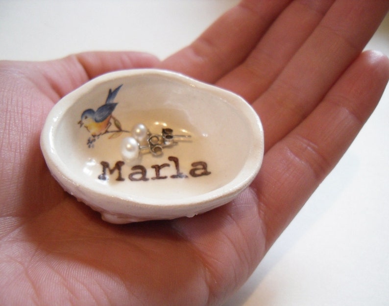 May include: A white ceramic dish shaped like a seashell with a bluebird design and the name "Marla" written on it. Two pearl earrings are inside the dish.