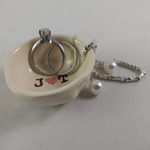 May include: A white ceramic ring dish with the initials "J &hearts; T" in brown lettering. The dish contains two silver rings, a silver necklace, and two pearl earrings.