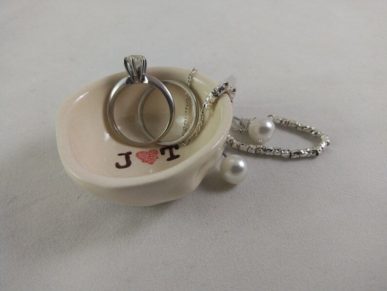 May include: A white ceramic ring dish with the initials "J &hearts; T" in brown lettering. The dish contains two silver rings, a silver necklace, and two pearl earrings.