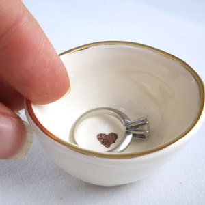 May include: A white ceramic ring dish with a gold rim and a small brown heart design. A silver ring with a diamond is sitting in the dish.