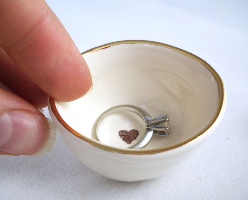 May include: A white ceramic ring dish with a gold rim and a small brown heart design. A silver ring with a diamond is sitting in the dish.
