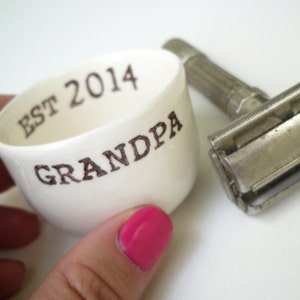 May include: White ceramic cup with the text "EST 2014 GRANDPA" in black lettering. A silver safety razor is next to the cup.
