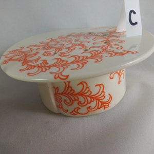May include: White ceramic cake stand with an orange floral design. The stand has a round top and a cylindrical base. The top has small holes for drainage.