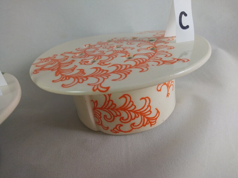 May include: White ceramic cake stand with an orange floral design. The stand has a round top and a cylindrical base. The top has small holes for drainage.