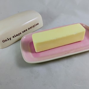 Puede incluir: Una mantequera con un plato rectangular rosa que contiene una barra de mantequilla amarilla. La tapa blanca tiene el texto marrón "Only slags use margine". Es un artículo de cocina.