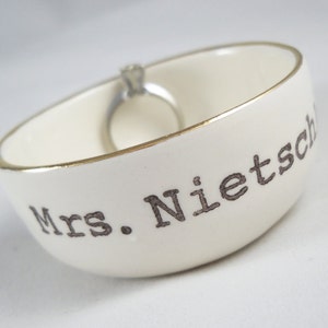 Puede incluir: Plato de cer&aacute;mica blanco para anillos con borde dorado y el texto "Mrs. Nietzsche" en letras negras.
