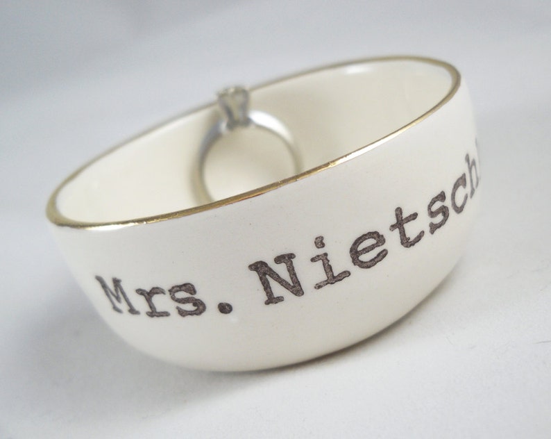 Puede incluir: Plato de cer&aacute;mica blanco para anillos con borde dorado y el texto "Mrs. Nietzsche" en letras negras.