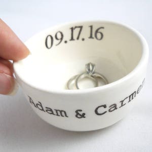 Puede incluir: Plato de cerámica blanco para anillos con el texto "Adam & Carmel" y la fecha "09.17.16" impresa en el lateral. Dos anillos de plata están dentro del plato.