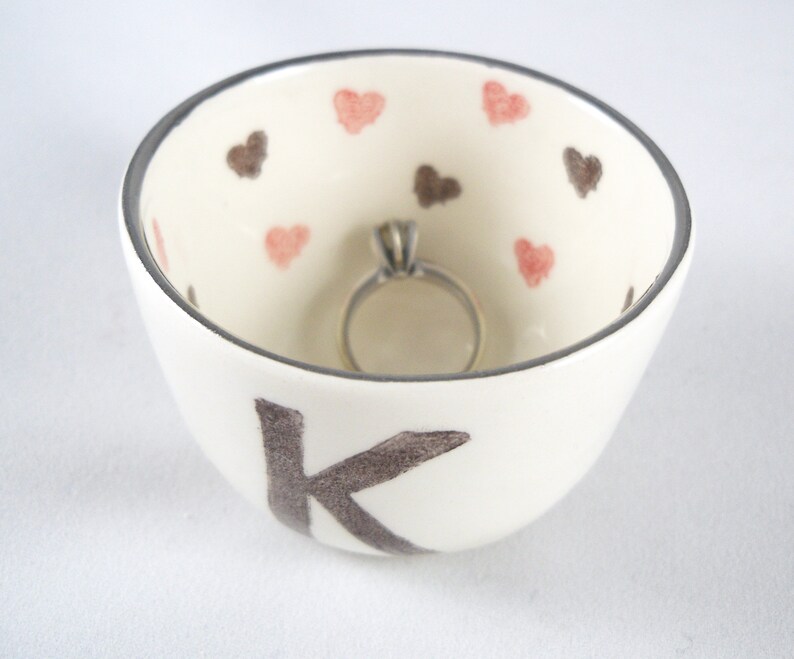 May include: White ceramic ring dish with a gray letter K and pink and brown hearts. A silver ring is sitting in the dish.
