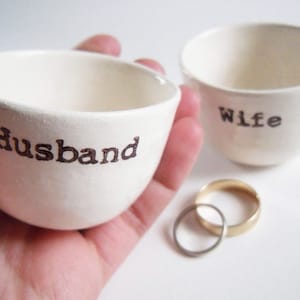 Puede incluir: Dos cuencos de cer&aacute;mica blanca con las palabras "Husband" y "Wife" escritas en ellos. Dos alianzas de oro est&aacute;n en el fondo.