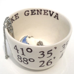 May include: Cream-colored ceramic bowl with the text "LAKE GENEVA" and coordinates. A silver chain hangs over the side. Inside the bowl are small, white, shell-shaped objects with blue and gray details.