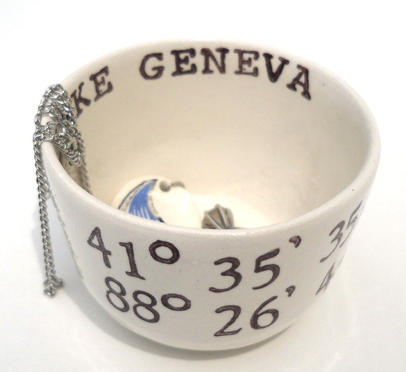 May include: Cream-colored ceramic bowl with the text "LAKE GENEVA" and coordinates. A silver chain hangs over the side. Inside the bowl are small, white, shell-shaped objects with blue and gray details.