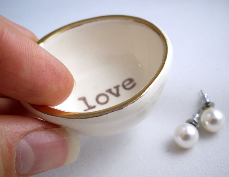 May include: A small white ceramic dish with a gold rim. The dish has the word "love" written on the inside. Two pearl earrings are next to the dish.