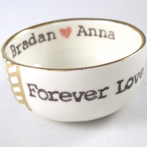 May include: White ceramic bowl with a gold rim and the words "Bradan" and "Anna" with a heart symbol between them. The words "Forever Love" are written on the bottom of the bowl.