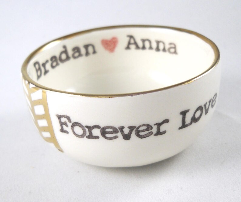 May include: White ceramic bowl with a gold rim and the words "Bradan" and "Anna" with a heart symbol between them. The words "Forever Love" are written on the bottom of the bowl.