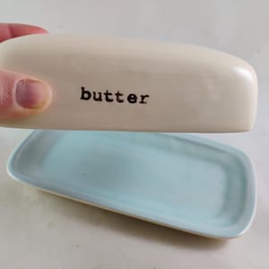 Op de afbeelding: Een keramische botervloot met een lichtbeige deksel en een lichtblauwe basis. Het rechthoekige deksel heeft het woord "butter" in zwart. De basis is rechthoekig met afgeronde hoeken.