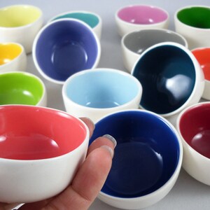 Puede incluir: Una colección de cuencos de cerámica coloridos con bordes blancos. Los cuencos están dispuestos en semicírculo con una mano sosteniendo uno de los cuencos en el centro.