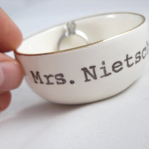 Puede incluir: Plato de cer&aacute;mica blanco para anillos con borde dorado y el texto "Mrs. Nietzsche" en negro.
