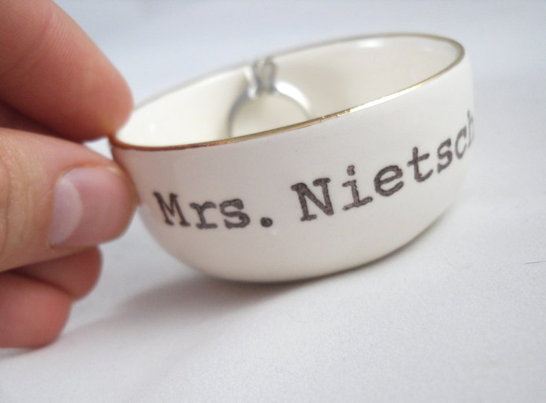 Puede incluir: Plato de cer&aacute;mica blanco para anillos con borde dorado y el texto "Mrs. Nietzsche" en negro.